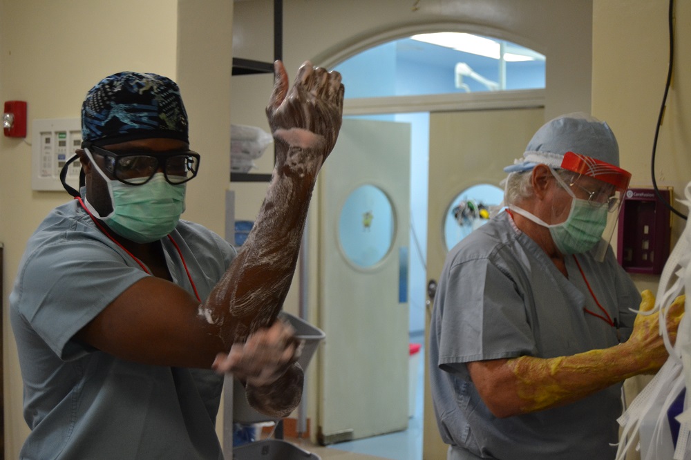 Two men in surgical scrubs stand beside each other, washing their hands and forearms to sanitize them before surgery. 