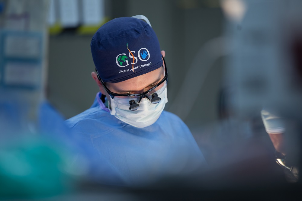 A close-up mage of a man's face, covered in a surgical mask, cap, and glasses, as he concentrates on a surgery. 